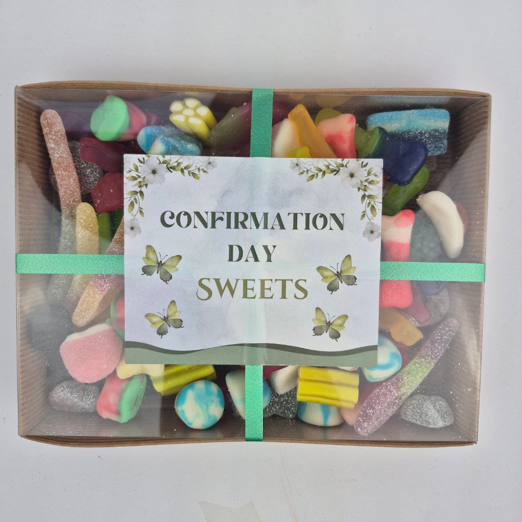 A clear gift box containing an assortment of colorful candies, sealed with a green ribbon and a label that reads 'CONFIRMATION DAY SWEETS'.