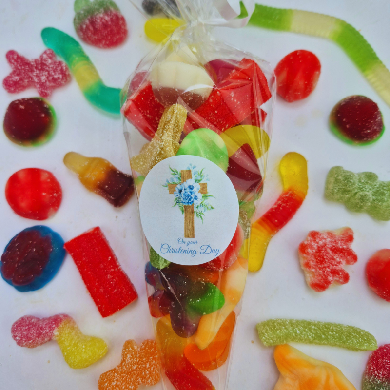 A clear cellophane gift bag filled with assorted colorful gummy candies and sour gummy worms, sealed with a white round sticker that reads 'On your Christening Day.' The bag is photographed on a white surface surrounded by scattered gummy candies.