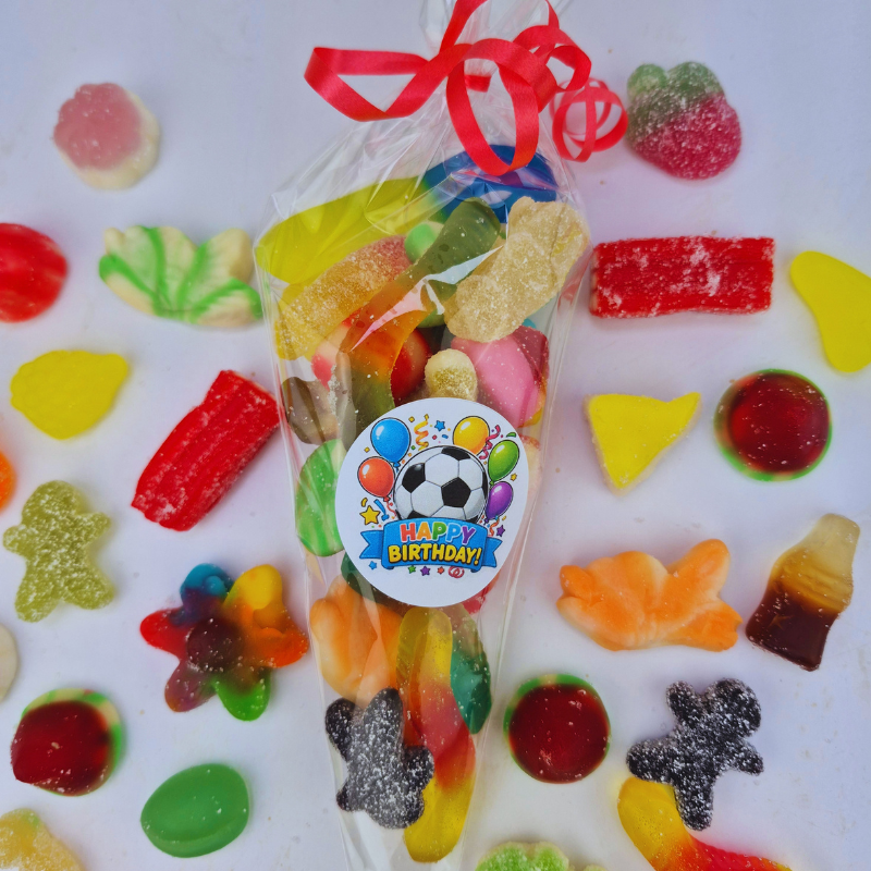 A clear cellophane bag filled with assorted colorful gummy candies and sweets, tied with a red ribbon and topped with a round 'Happy Birthday' sticker. The bag is photographed on a white background surrounded by scattered gummy candies.