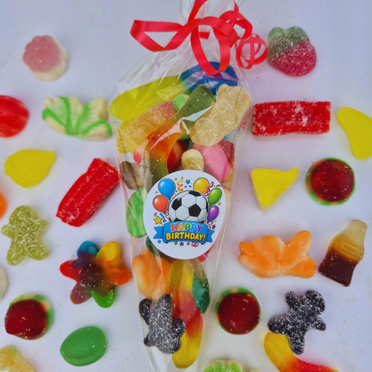 A clear cellophane bag filled with assorted colorful gummy candies and sweets, tied with a red ribbon and topped with a round 'Happy Birthday' sticker. The bag is photographed on a white background surrounded by scattered gummy candies.